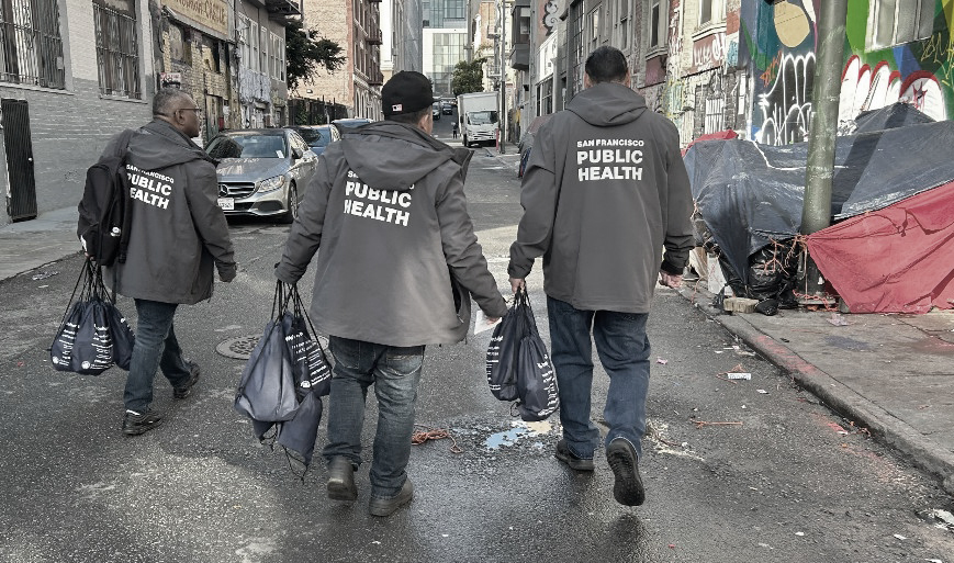 Three San Francisco Public Health workers with care kits walking toward a tent on the street